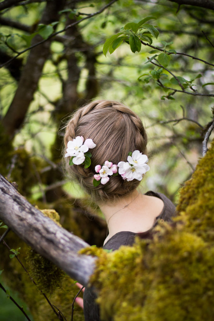 A girl with pink and white apple blossoms in her golden braid, gracefully tilts her head to look through the rich mossy branches in which she is perched.